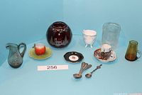 Full lot photo showing all glassware and ceramic items including the red etched crystal bowl, clear jar, small pitchers, souvenir cup and saucer, black dish, and spoons.