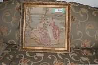 Framed tapestry showing two ladies in period dresses in a pastoral scene, sitting on a patterned couch.