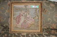 Full view of framed tapestry of two ladies in pink dresses outdoors with scenery background on a couch with floral upholstery.