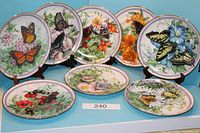 Eight decorative round plates featuring different butterfly and floral designs arranged with five standing on plate holders and three lying flat, clearly showing colorful and detailed butterfly illustrations.