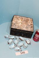 Black tin container filled with clear crystal prisms, plus several prisms displayed outside tin next to a soda can for scale.
