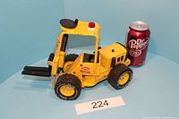 Yellow 1970's Tonka forklift toy shown from left side with black adjustable forks extended, large black tires with yellow rims, and red light on top. Dr Pepper can in background for size comparison.