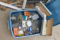 Wide view of tote filled with assorted hardware items including boxes of electrical outlets, small plastic containers with fasteners, and other materials alongside tools and chicken wire roll in background.