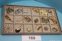 Photo shows a tray with 18 assorted brooches laid out in 4 rows. Designs include butterflies, flowers, a dog, birds, and abstract shapes in gold and silver tones. Some brooches are embellished with rhinestones and faux pearls.