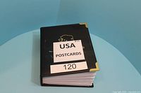 Closed black album with gold corner protectors labeled 'USA POSTCARDS 120'
