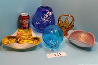 Five art glass items including blue glass cat, yellow art glass bowl, blue vase, orange crown-like sculpture, and pink bowl shown with Dr Pepper can for scale