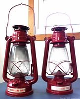 Front view of two red American Camper RK12 kerosene lanterns with glass chimneys and metal guards.
