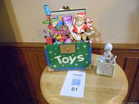 Precious Moments music box with a green box labeled 'Toys' and a Christmas-themed teddy bear figurine with toys on top, placed on a round wooden stool.