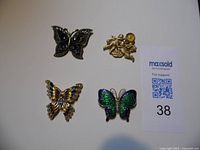 Four vintage brooches displayed on white background with a MaxSold lot number card showing number 38. Three butterfly brooches and one cherub brooch with rhinestone details visible.