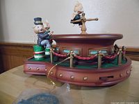 Side view of vintage Enesco music box showing three pig figurines on a circular platform with turntable figurine in the center and decorative rope with gold-tone accents.