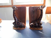 Front view showing paired giraffe carved bookends with detailed giraffe neck and head attached to a flat wood backing, placed on blue surface near window.