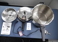Top down view of the stainless steel pot, lid with detachable handle and pressure regulator, two perforated steaming inserts, instruction manual, and additional components.