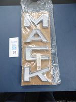 Front view of four 'MACK' letters mounted on cardboard, sealed in plastic bag