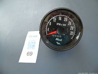 Front view of round analog vintage Mack tachometer gauge with black dial, white numbers for RPM up to 3200, red needle, and Mack logo with odometer window.