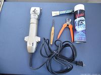 Oster-Stewart Clipmaster Model 510A electric hair clipper with attached cord, and accessories including tube of clipper grease, wooden brush, orange blade remover, and lubricant spray can arranged on blue surface.