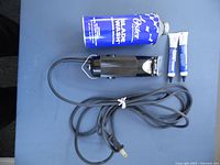 Black Oster Golden A5 clipper with attached power cord, blue can of blade oil, and two tubes of lubricant on a blue surface.