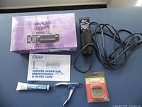Photo showing the Oster Single Dog Clipper with original purple box, instruction manual, clipper grease tube, dog nail clipper, extra clipper blade in package, and clipper oil bottle all arranged on a gray surface.