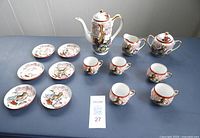 Full set arranged on a blue surface showing all pieces including teapot, sugar bowl, creamer, tea cups, and small plates with intricate peacock design.