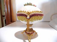 Front view showing white egg with floral embossing, pink rhinestones, faux pearls, and gold-tone trim on ornate tripod base.