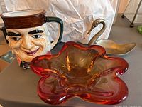 Photo showing the art glass flower dish with deep red and amber hues, the ceramic Toby jug mug with detailed male face wearing a brown hat, and the silver-toned metal swan figurine.