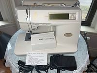 Front view of Babylock Ellure computer sewing machine with Pfaff Hobbylock 4752 cover, foot pedal, power cord, and handwritten note with details.