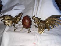 Two brass rooster figurines with a reddish alabaster egg between them on a white cloth background. The rooster on the left has one leg broken off, the alabaster egg sits on a tripod stand.