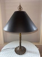 Full view of the Chapmans brass table lamp with black lampshade sitting on a white lace doily.