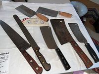 Photo of eight butcher knives and cleavers placed on white cloth. Shows variety in size and handle types including wooden and black.