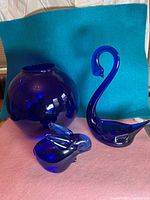 Three cobalt blue glass pieces including rabbit figurine, swan figurine, and vase against blue and pink fabric background
