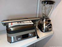 Cuisinart Panini Press closed with digital control panel and Oster blender with glass pitcher, both placed on a white countertop.