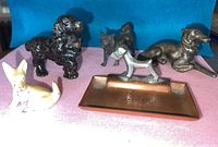 Photo showing five miniature metal dog figurines including a black cocker spaniel, grey Afghan dog, small terrier, cream-colored lying dog, and silver-grey Art Deco greyhound on a brass ashtray.