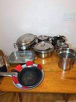 Full view of various pots, lids, roasting pan, steamer, and strainers arranged on a table with cast iron and non-stick pans in front.