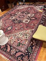 Full view image of rectangular wool rug with geometric and floral patterns, multi-color including pink, navy, green, and cream, with fringe along the short edge. Shows scale in a room setting.