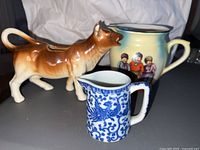 Photo showing three vintage ceramic creamers: a brown and white cow-shaped creamer, a round ceramic creamer with painted figures, and a smaller blue and white patterned creamer.