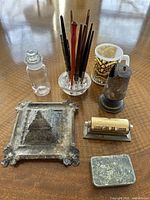 Vintage desk items arranged on wood table including glass ink well with wooden pens, metal tin with nibs, brass calendar, silver-tone tray, match holder and Fire King mug.