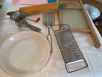 Photo showing vintage kitchen items: glass washboard, wooden rolling pin, metal hand-crank meat grinder, white pie plate, metal grater, and wire basket.
