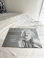 Metal tin sign with black and white photo of Marilyn Monroe with a quote and autograph on a table with white lace tablecloth.