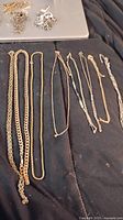 Photo of eight gold tone chains laid out on a fabric surface, showing various styles and lengths.