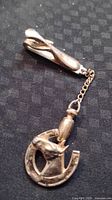 Close-up photo of the silver-tone Hickok tie clip with horse head inside a horseshoe charm hanging from a chain.