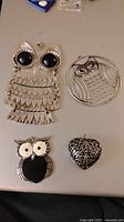 Four silver tone pendants laid out on a surface including three owl designs and one heart
