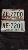 Front view of two 1973 Colorado license plates with the registration AE-7200 placed one above the other on a dark fabric background.