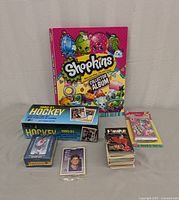 Overview of collection showing Shopkins collector album, sealed Shopkins card pack, 1990-91 hockey card box, Powerplay black card deck, and assorted cards.