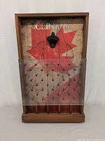 Front view of Clink It game board showing red maple leaf design, metal bottle opener, peg holes protected by clear plastic, and wooden bottle cap compartments