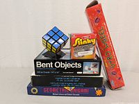 Photo of all items including Rubik's Cube on stand, Slinky toy box, Gyro Wheel box, Bent Objects puzzle, metal brain drains puzzle, and origami books.