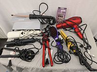 Overall view of hair care and beauty items including hair dryer, curling irons, straighteners, hair brush, and pack of false nails, cords mostly tangled, displayed on gray surface.