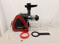 Photo of Homever electric juicer in black and red, including juicing and pulp containers, red sealing ring, and black tool, placed on a flat surface with a white background.
