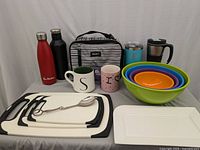 Lot overview showing lunch bag, water bottles, mugs, cutting boards, mixing bowls, and plates on white backdrop