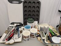 Photo showing white-handled cutting board, black mini muffin tray, assorted mugs, metal tray, placemats, and assorted knives with colorful handles