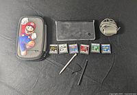 Photo showing the Nintendo DS console, Mario case, six game cartridges, AC adapter, two styluses, and game card holder on a black surface.