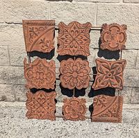 Front view of brown metal wall decoration with nine distinct tile designs featuring floral and geometric motifs against a concrete block wall.